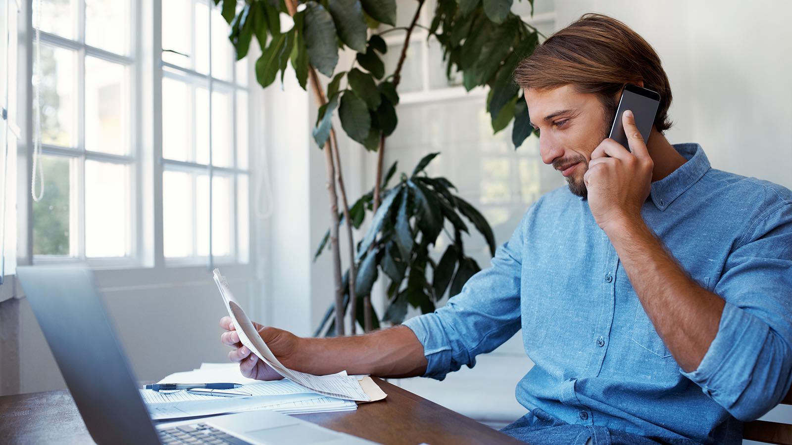 Man at desk using phone reviewing documents.