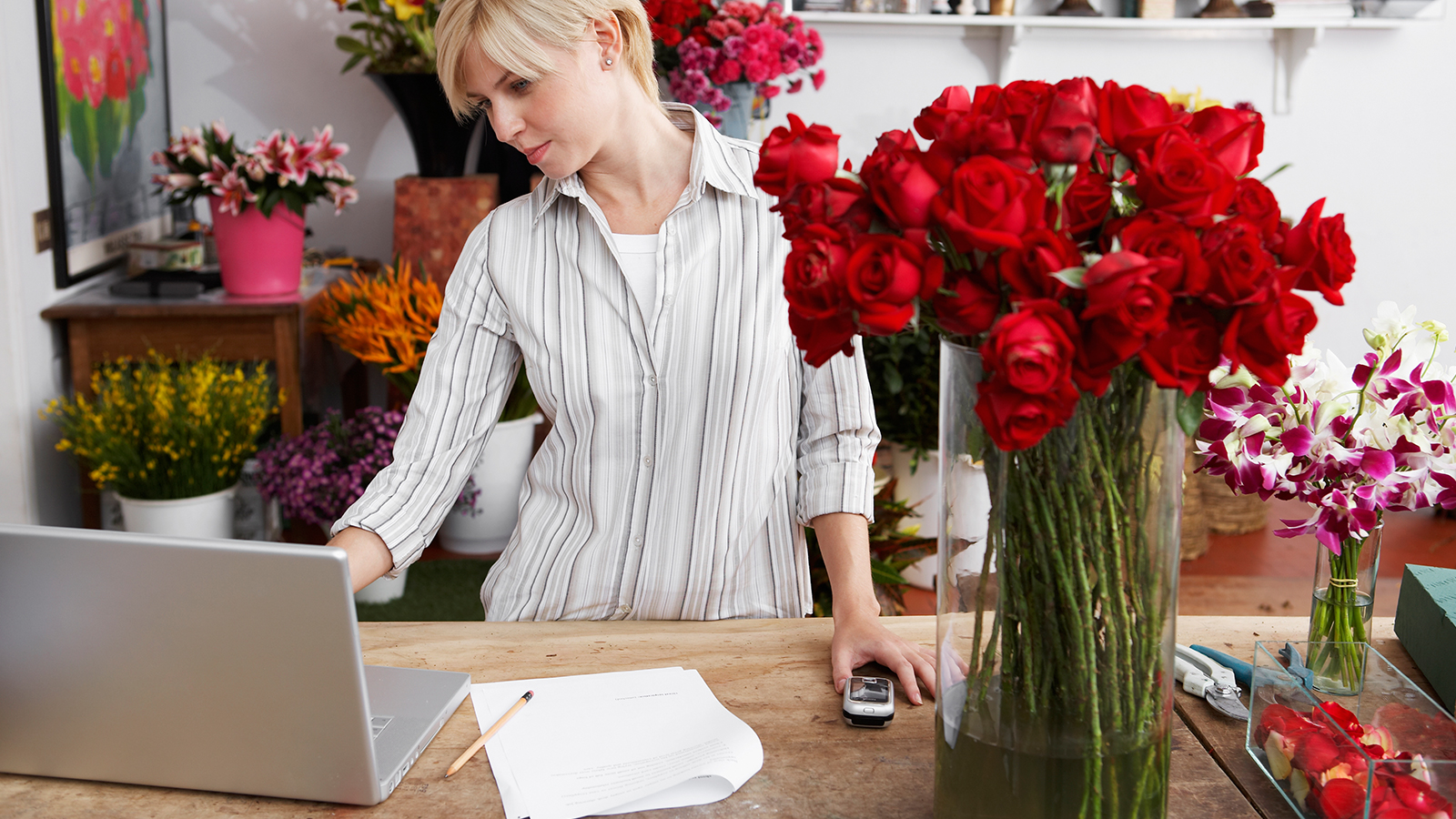 A woman standing at a table inside a flower shop, looking at her laptop.