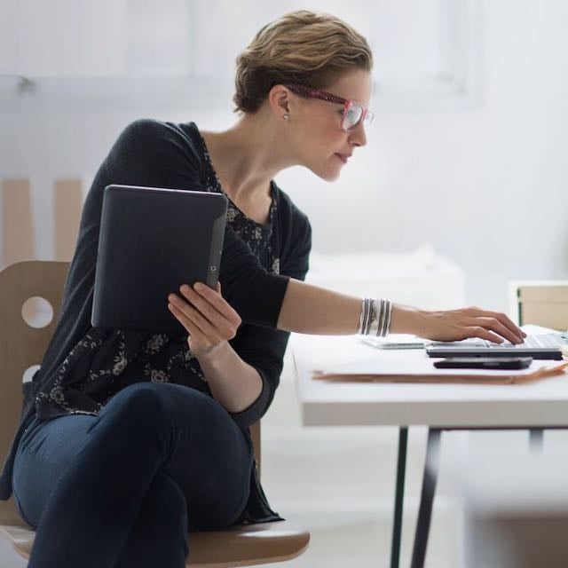 Woman sitting at a desk, holding a tablet and typing on a laptop.