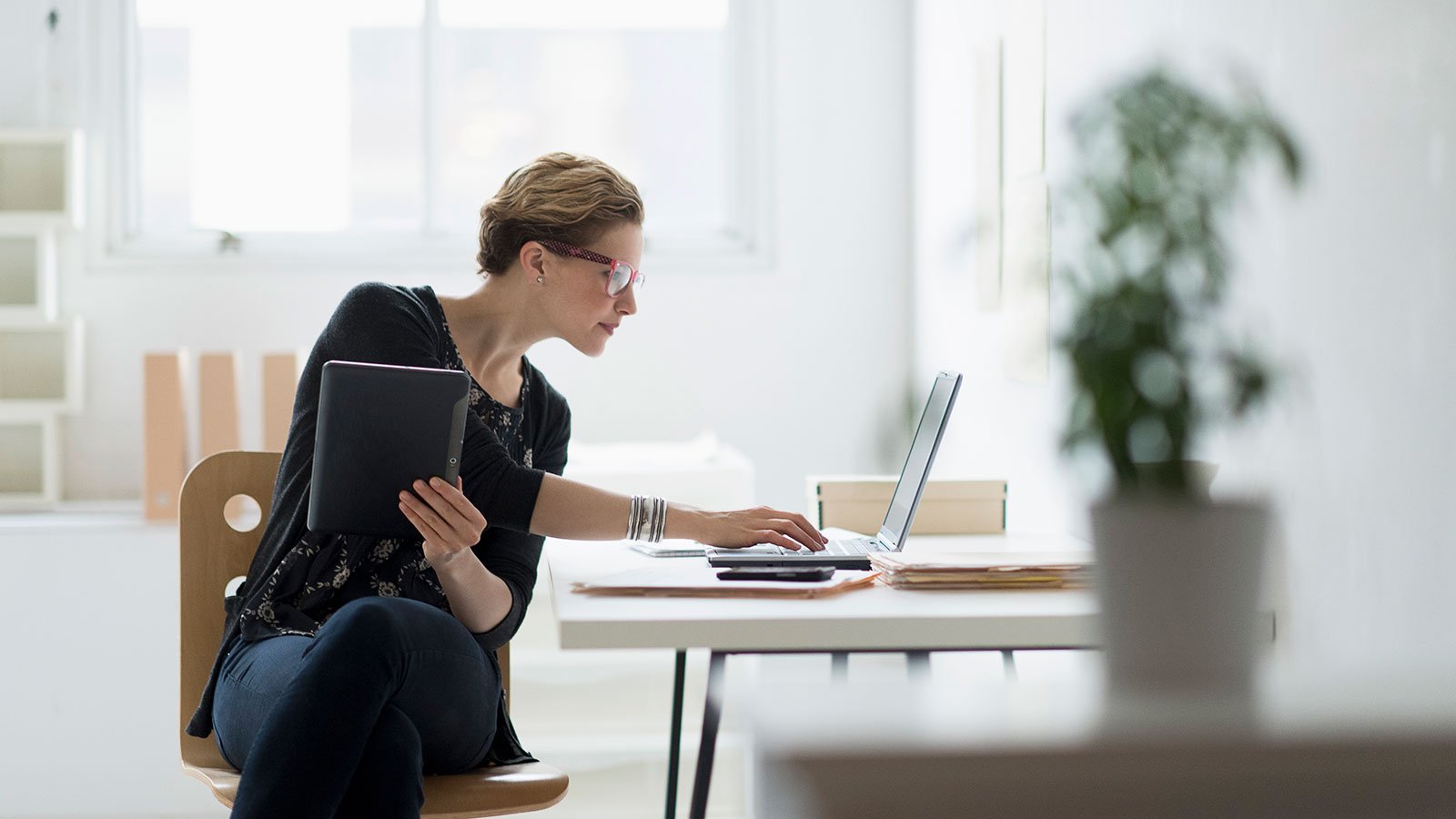 Woman sitting at a desk, holding a tablet and typing on a laptop.