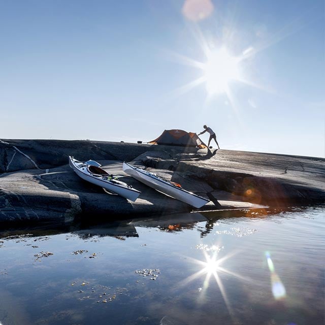 Two kayaks on the shore and a man setting up a tent nearby.