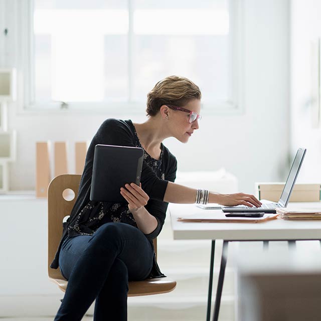 Une femme assise au bureau, tenant une tablette, et tapant sur un ordinateur portable.